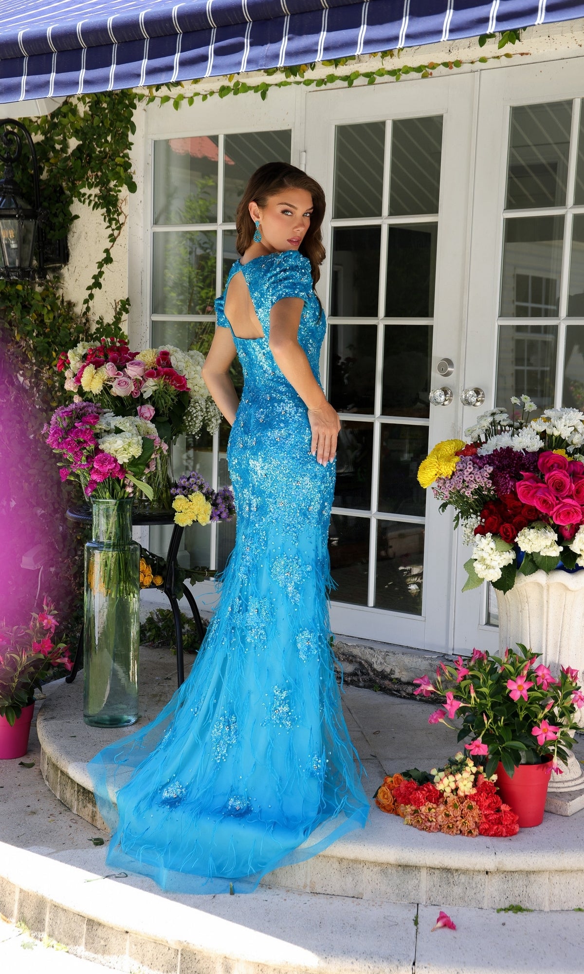 A woman wears the Feather Short-Sleeve Prom Gown: Ava Presley 39552, standing on a patio with flowers and glass doors, surrounded by vibrant floral arrangements.